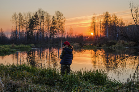 Woman catches a fish on spinning  In calm weather at sunset, she is standing on the shore of the pond.の写真素材