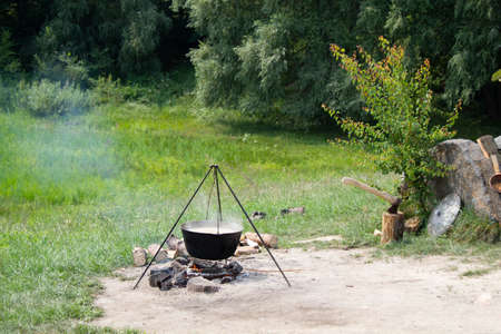 Old cast-iron cauldron on a fire. Cooking food outdoors in a pot on a tripod. Ax in a wooden stump....