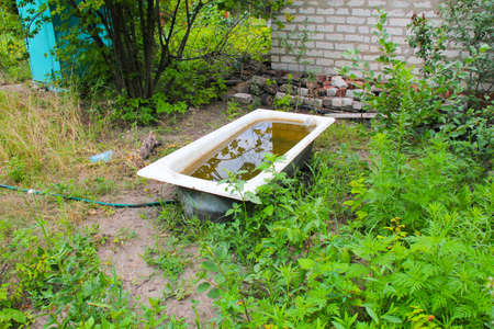 Bath with dirty water in the garden. Ecology and nature pollution concept. Ecological catastrophe. Bathtub with rusty water in green grass next to a brick house and a wooden toiletの写真素材