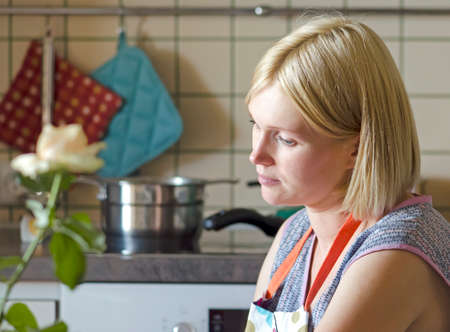 Young intelligent pretty housewife sitting at table. Looks thoughtfully at the screen. On the face of sadness and confusion. Preparing food in the comfortable modern kitchen.の写真素材
