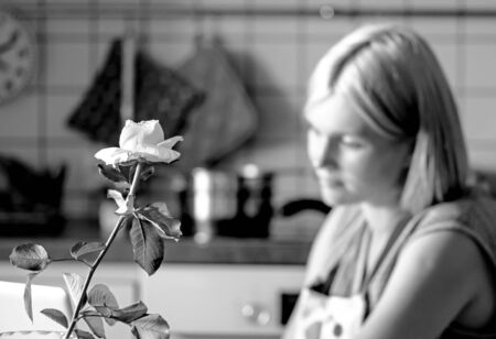 Young intelligent pretty housewife sitting at table. With interest read the recipe on screen. In front of her is a vase with rose. Comfortable modern kitchen. Only flower is in focus. Black and White.の写真素材