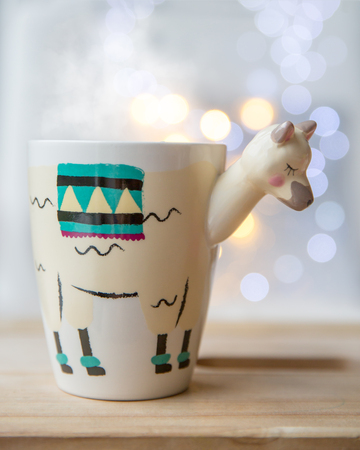 Beautiful llama shaped trendy cup with hot tea with lights on background. Cozy autumn-winter conceptの写真素材