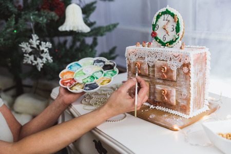 Women decorates ginger cookies Christmas chest of drawers at home. Woman draws paints on honey gingerbread cookies. The chest of drawers is made of gingerbread and decorated with clock and small figures of bears. Preparing gifts for loved onesの写真素材