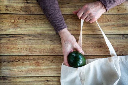 Hands of an old woman put fresh organic avocado in Eco Shopping Bag on wooden background, Flat Lay. Top View. Ecology protection conceptの写真素材