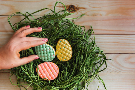 Woman's hand takes one of handmade colorful easter eggs cookies from nest on light wooden background