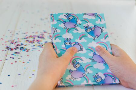 Hands of a girl holding notebook with llama unicorn and rainbow on white wooden background with...