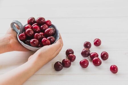 Kids hands with fresh cherries in light blue plate on white wooden background. Summer and harvest conceptの写真素材
