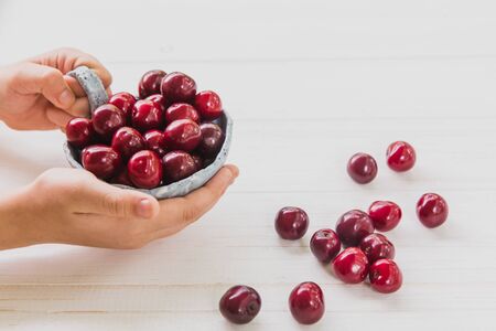 Kids hands with fresh cherries in light blue plate on white wooden background. Summer and harvest conceptの写真素材