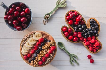 Acai smoothie, granola, seeds, fresh fruits in a wooden bowl with cactus spoon. Plate filled with berries on white wooden background with with Monstera leaf.の写真素材