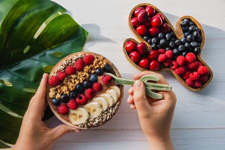 Acai smoothie, granola, seeds, fresh fruits in a wooden bowl in female hands with cactus spoon. Plate filled with berries on white wooden background with with Monstera leaf.の写真素材