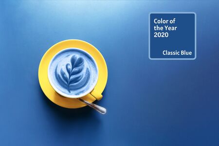 Yellow cup with latte in blue color with art flower petals on the foam on blue table background. 2020 color trendtextの写真素材