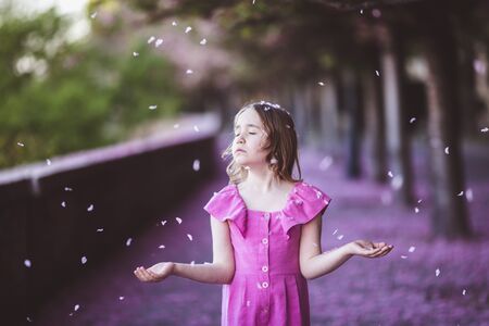 Beautiful girl in pink dress in cherry blossom park on a spring day, flower petals falling from the...