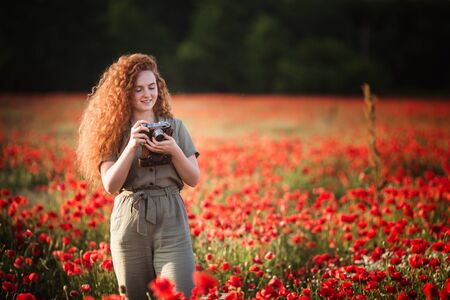 Young beautiful red-hear girl takes photos in the red poppy flowers field on sunset
