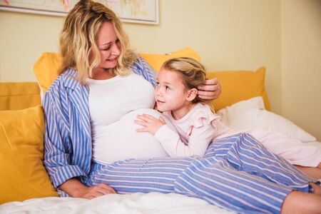 Pregnant woman in blue striped pajamas and her daughter are happy and hugging in bedroom in the...
