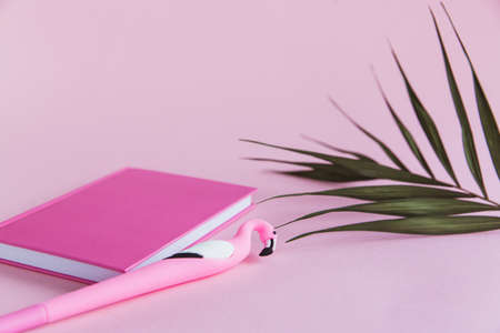 Pink notebook for notes, funny flamingo pen and green palm leaves on pink pastel backgroundの写真素材