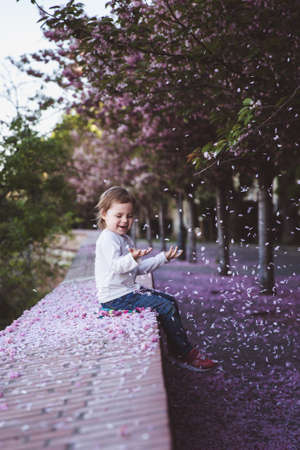 Beautiful girl 5 y.o. sit in cherry blossom park on a spring day, flower petals falling from the treeの写真素材
