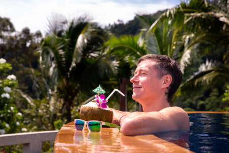 Handsome man relaxes by the outdoor pool and drinks a coconut with palm trees in the background. Summer concept.の写真素材