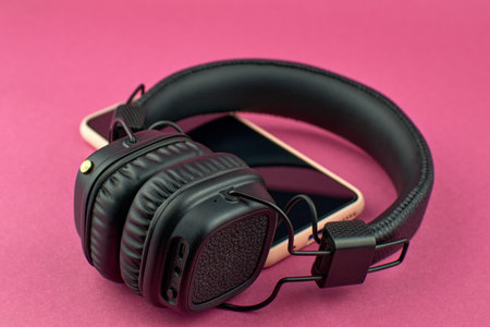 Wireless black headphones and smartphone. Black over-ear headphones on pink background, soft focus.