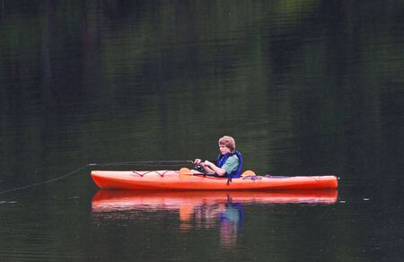 A young teenage boy fishing from a kayak in the evening. It is raining and streaks of rain can be...