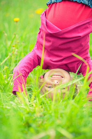 Happy little girl standing upside down on green grass in summer parkの写真素材