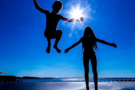 Silhouette of two kids jumping on trampoline against blue skyの写真素材