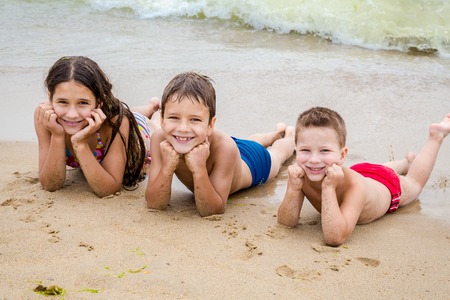 Three smiling kids on the beach lying down on the sand near waterの写真素材