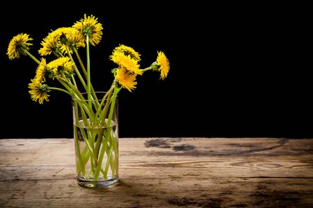 Bouquet of yellow dandelions in glass on grungy wooden table and black background with empty space for textの写真素材