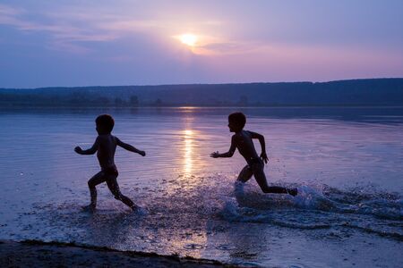 Silhouette of two boys running on river's beach against sunsetの写真素材