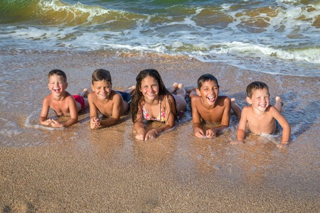 Five smiling kids on the beach lying down on the sand near waterの写真素材