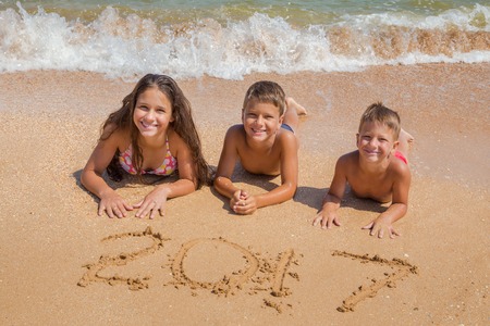Three smiling kids on the beach lying down on the sand near water with new year 2017 sign