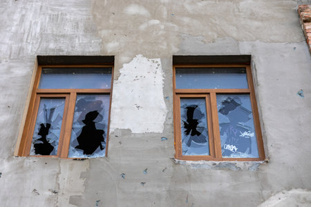 Two damaged wooden-framed windows with shattered glass on a weathered concrete wall, symbolizing...