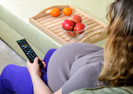 Pregnant woman watching television holding a remote control at homeの写真素材