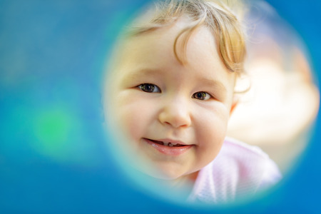 The one-year child plays on the playground. Cute baby girl looks through a round window to the camera.の写真素材