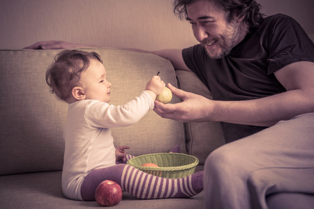Happy baby girl plays with fruits with her father at home. The one-year child holds a pear in hand.の写真素材