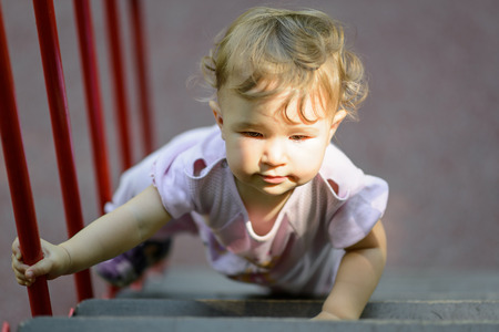 The one-year child climbs up the stairs. Cute baby girl plays on the playground.の写真素材