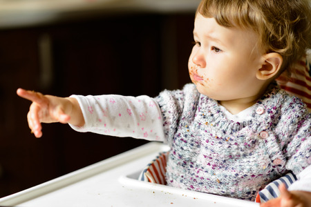 Eating baby girl with messy face and full mouth of food. The one-year child points his finger, demanding more food.の写真素材