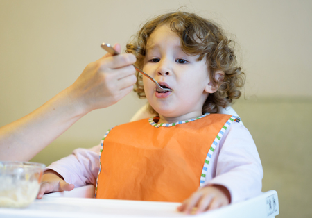 The two-year child eats porridge and looks TV. Little girl opens mouth to eat.の写真素材
