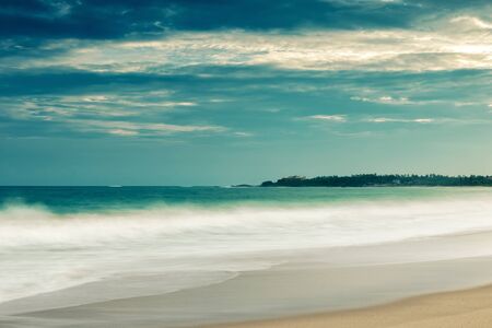 Sandy tropical beach with long exposure for blurred water, milky wave effect. Sea beach background. Panoramic view of the ocean coast and the dramatic sky. Beach with surf in twilight.の写真素材