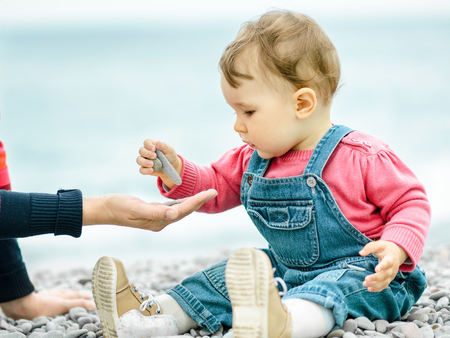 Beautiful baby with mother play on the beach. One-year-old child puts a small stones in mom hand. Baby girl in denim overall sits near water edge. Cute toddler is outdoor in spring or autumn close-up.の写真素材