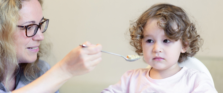 Baby girl during eating at home. Mother feeds her cute child with a spoon. Adorable toddler sits on high chair with food. The two-year-old kid eats porridge indoor. Panorama of family in kitchen.の写真素材