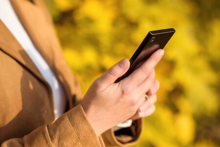 Woman using mobile phone outside. Hands holding cellphone close-up. Adult girl looks at black smartphone in autumn park. Person watching cell phone on street on sunny fall day. Technology concept.の写真素材