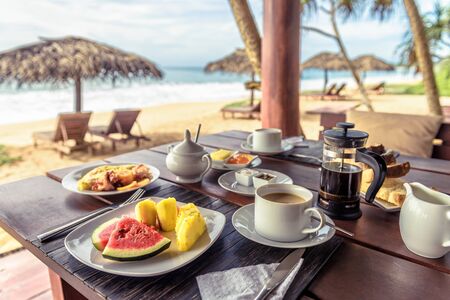 Breakfast on sea beach in Sri Lanka. Table setting with vegan food and coffee in restaurant outdoor. Buffet with exotic fruits on ocean coast. Concept of travel and vacation in tropical resort.の写真素材