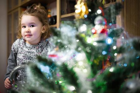 Baby girl hangs up decorations on Christmas tree at home. Cute little child is eve the Christmas and...