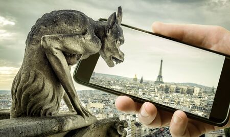 Gargoyle on Cathedral of Notre Dame de Paris looks at Eiffel Tower in mobile or cell phone, Paris,...