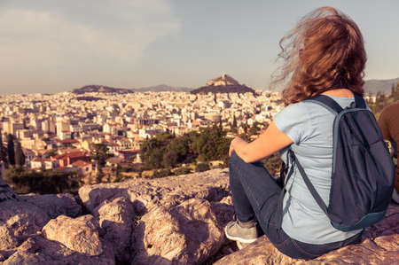 Young person on background of urban landscape of Athens, Greece, Europe. Adult pretty girl tourist relaxes on hilltop overlooking Athens city in summer. Concept of adventure and travel in Athens.の写真素材