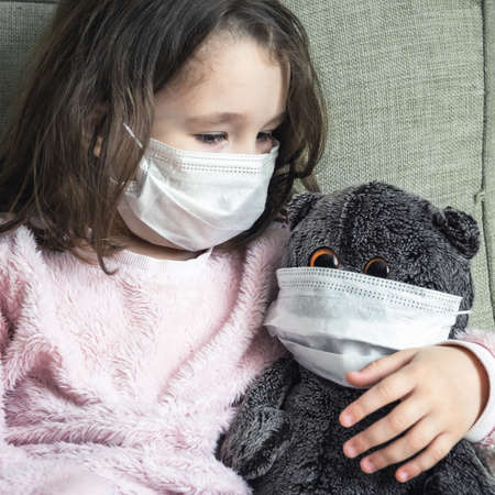 Kid in mask playing with soft toy during COVID-19 coronavirus pandemic. Cute little girl sits on couch indoor due to quarantine. Concept of safety, child protection, corona virus and restrictions.の写真素材