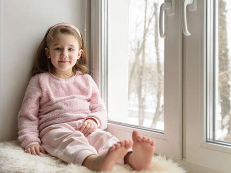 Kid sitting by window looks at camera indoor, portrait of pretty little girl on fur rug on room sill in winter. Adorable smiling child in pink is on windowsill in white interior at home.の写真素材