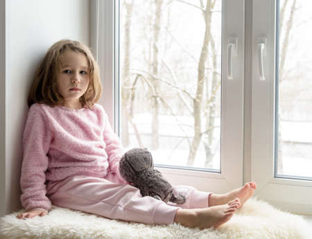 Kid sits on windowsill at home and looks at camera, portrait of pretty little girl on fur rug on room sill in winter. Adorable pensive child in pink, cute sad toddler with toy in white interior.の写真素材