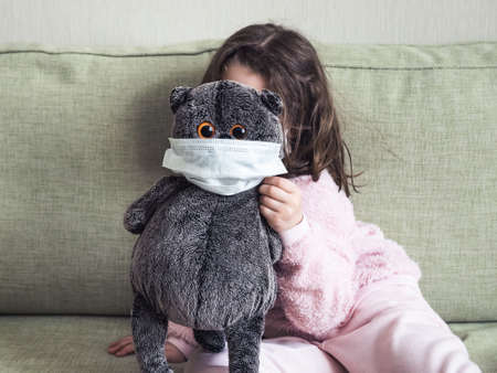 Kid playing with soft toy during COVID-19 coronavirus pandemic, funny playful little girl hides behind masked cat indoor due to quarantine. Concept of safety, child protection and corona virus.の写真素材