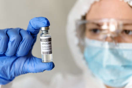 COVID-19 vaccine in doctor's hand close-up, nurse in medical PPE suit holds bottle with vaccine for coronavirus. Concept of corona virus treatment and vaccine research during pandemic.の写真素材